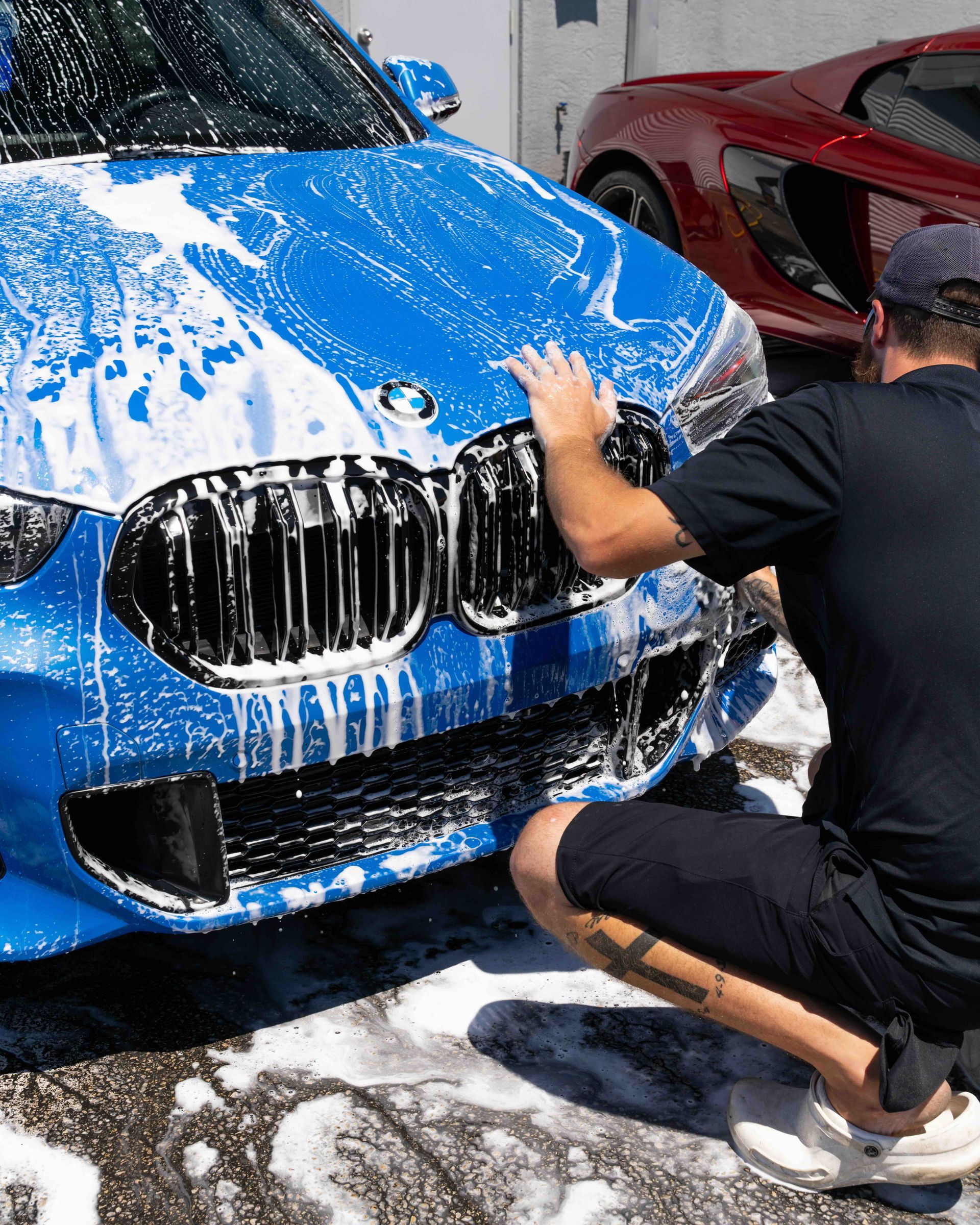 A man is kneeling down to wash a blue car.