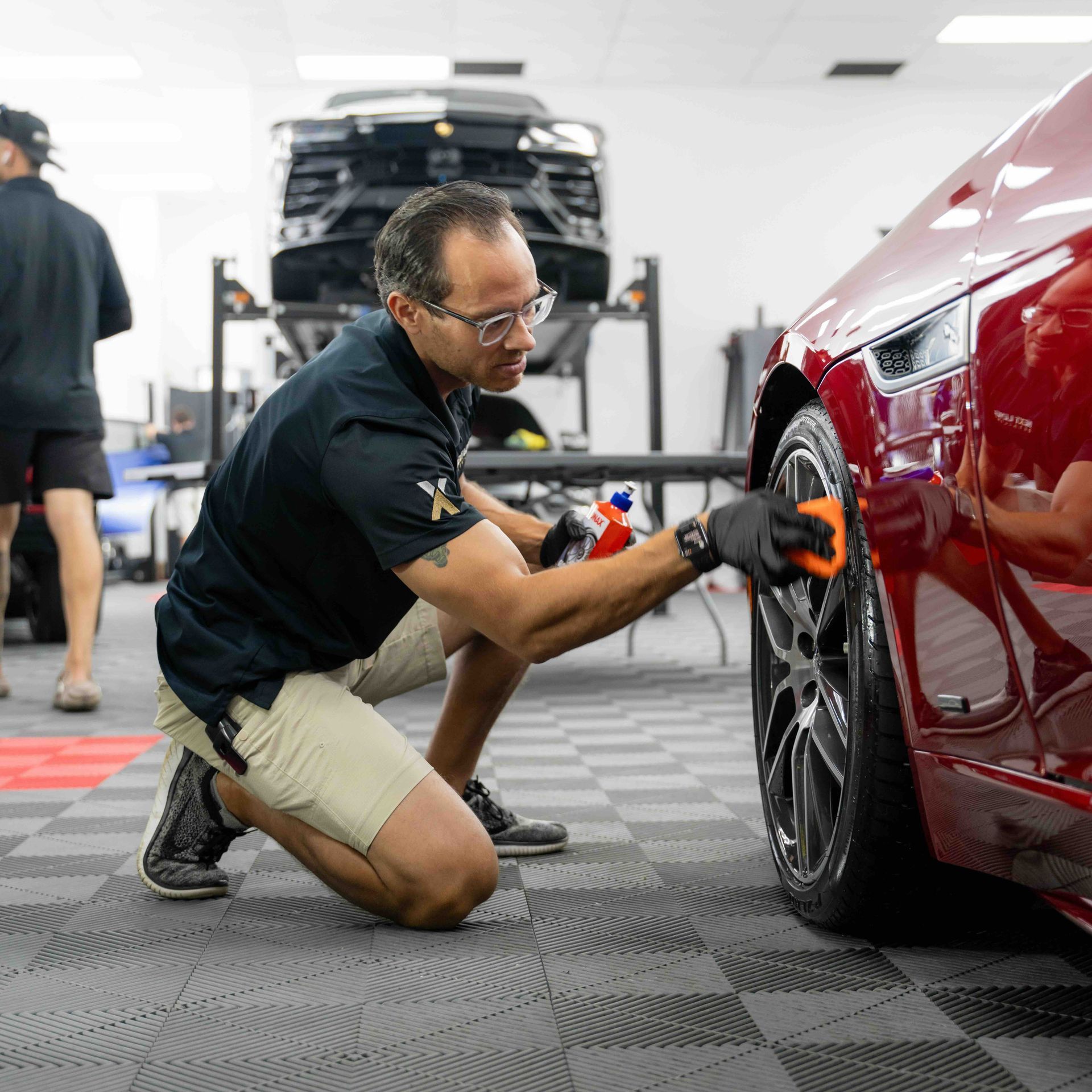A man is kneeling down to polish the wheel of a red car.