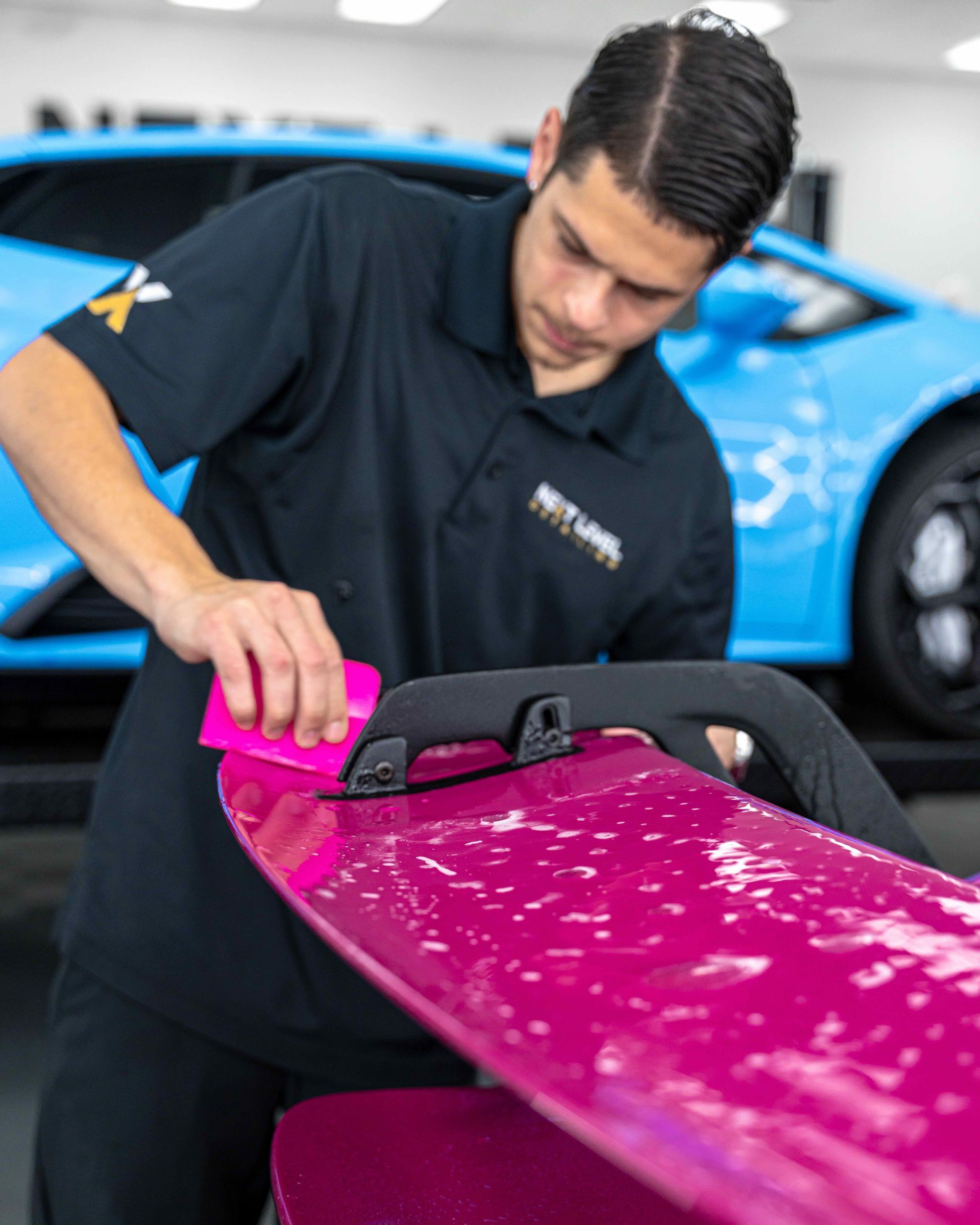 A man is polishing a pink surfboard in front of a blue car.