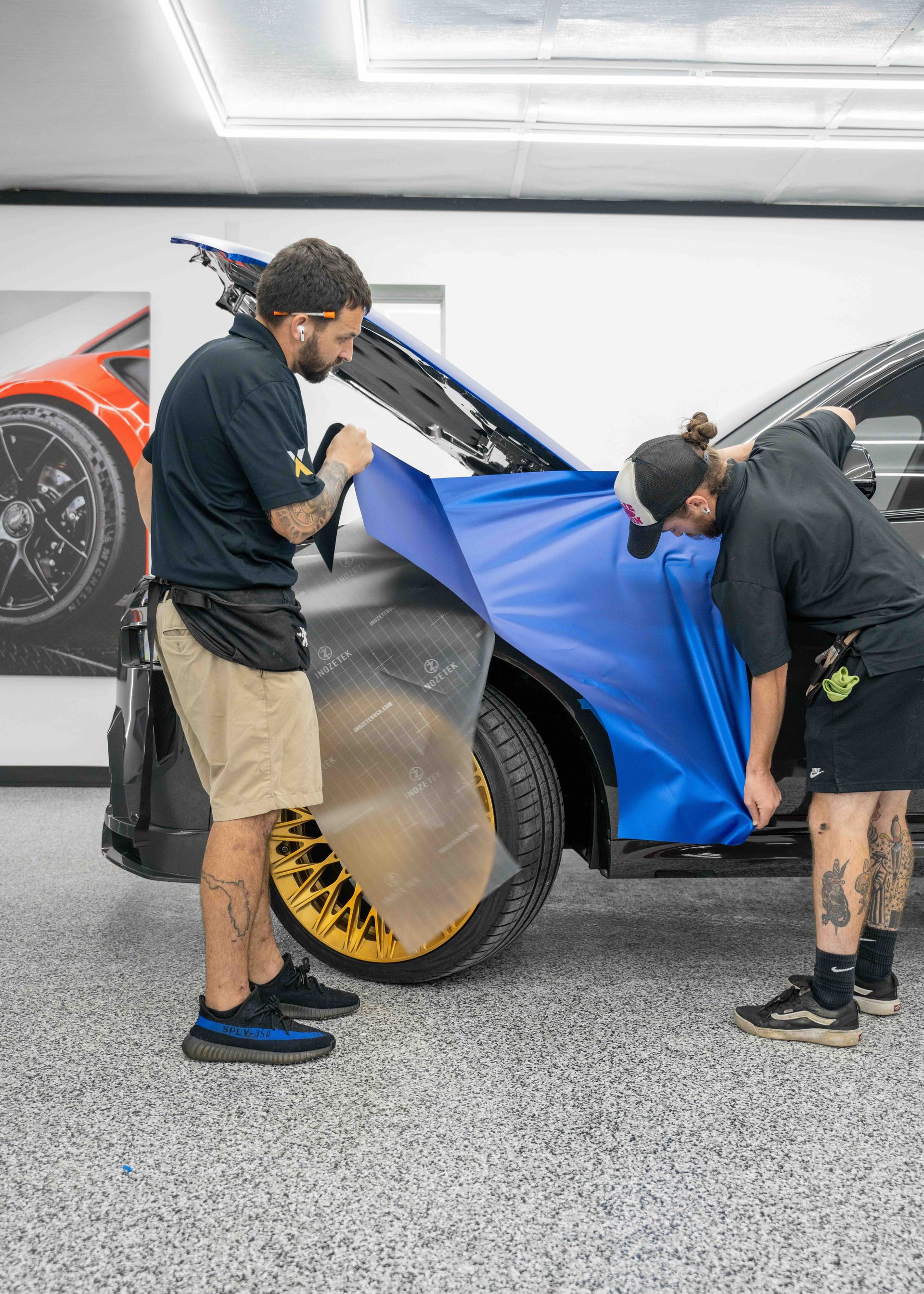 Two men are wrapping a blue car in a garage.