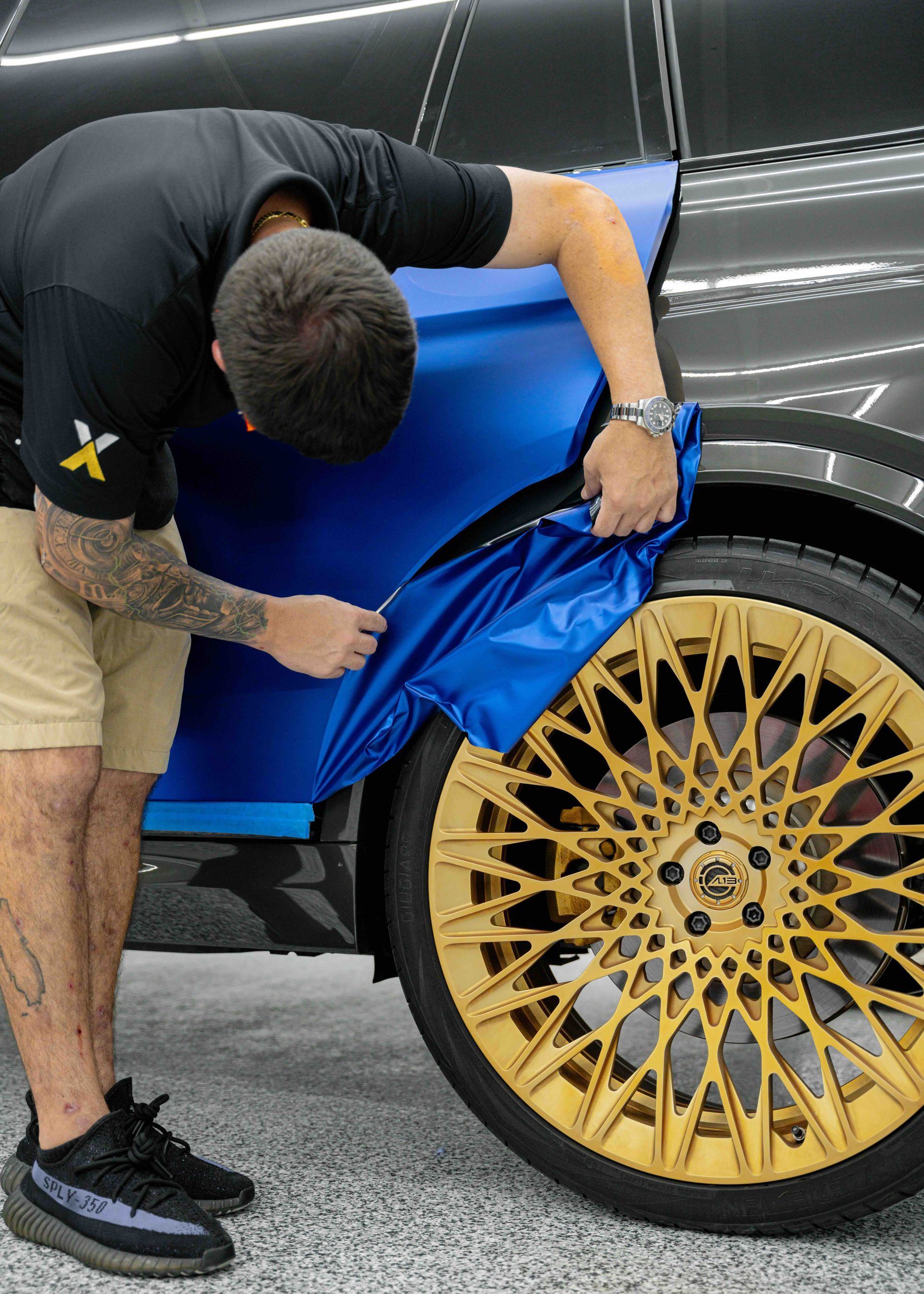A man is wrapping a blue car with gold wheels.