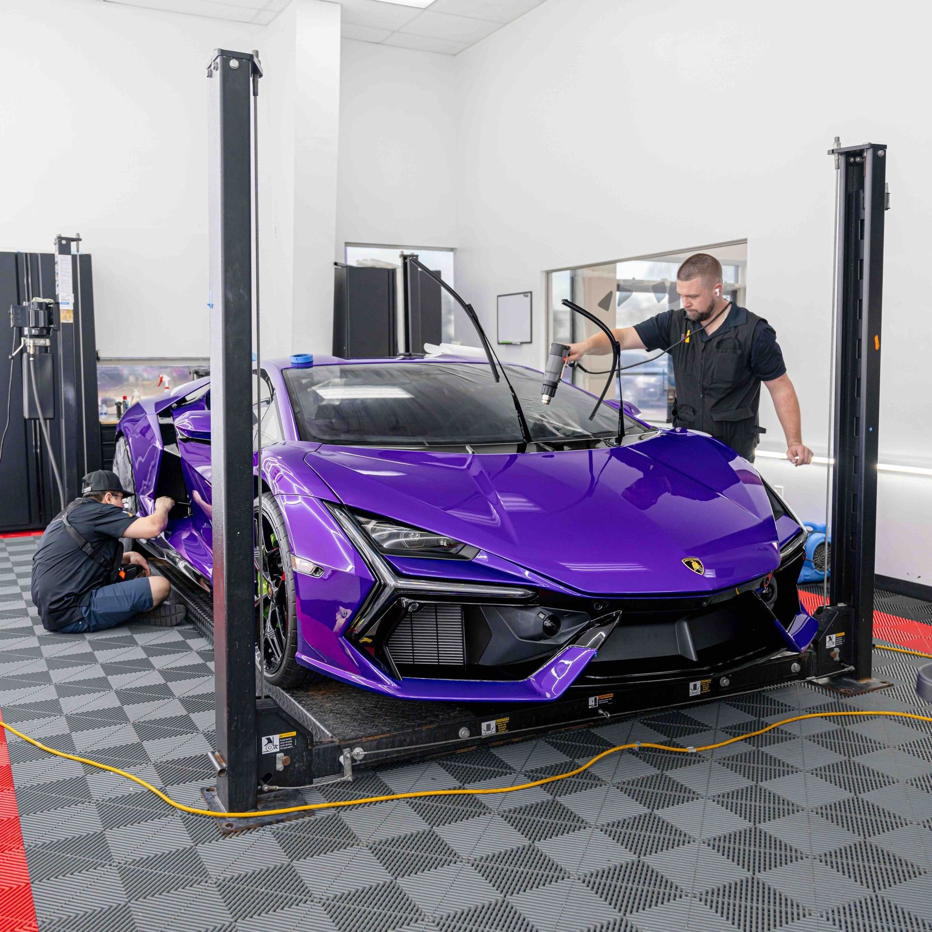 A man is working on a purple sports car on a lift in a garage.