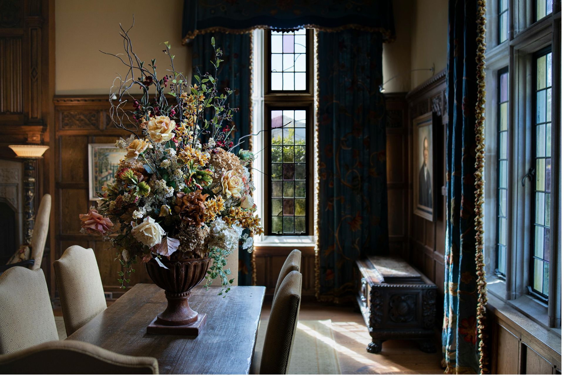 Formal dining room with a long wooden table, large flower arrangement, and ornate dark paneling. Sunlight streams through a window.