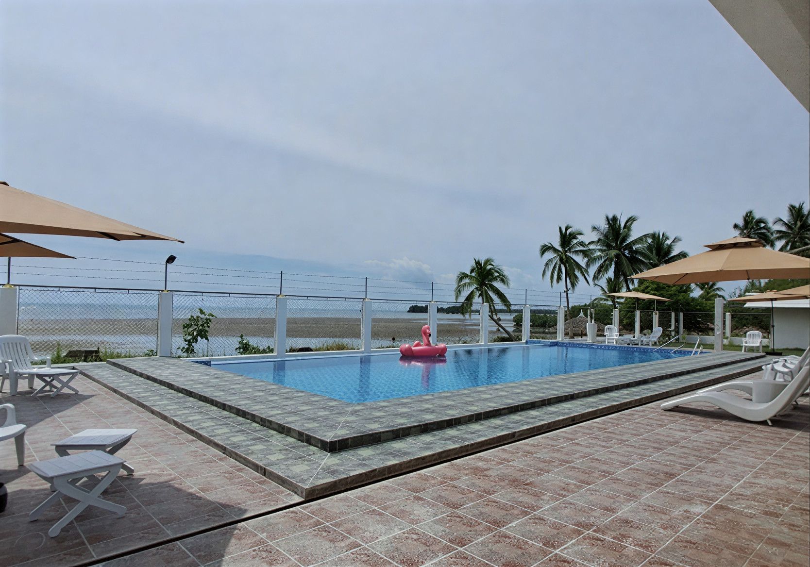 Swimming pool overlooking a beach, with palm trees and umbrellas. A pink flamingo float is in the pool.