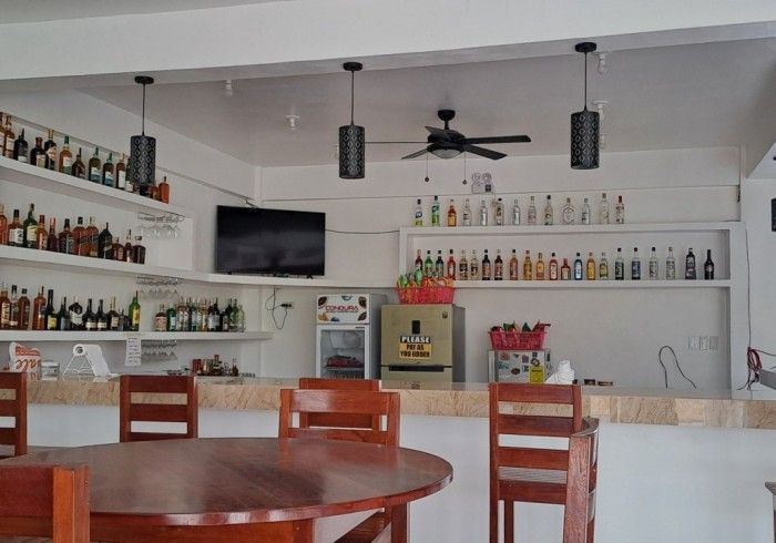 Bar interior with shelves of bottles, overhead lamps, a TV, and round wooden tables.