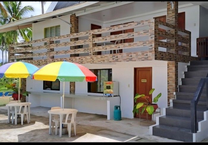 Exterior of a two-story building with a brown door, tables, and colorful umbrellas. Concrete stairs.