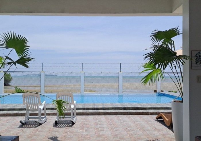 Two white lounge chairs beside a pool overlooking a beach. Blue sky, water, and palm trees.