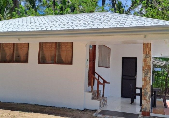White-walled bungalow with gray roof, windows, and brown door. Porch with stairs, tile, and brown columns.