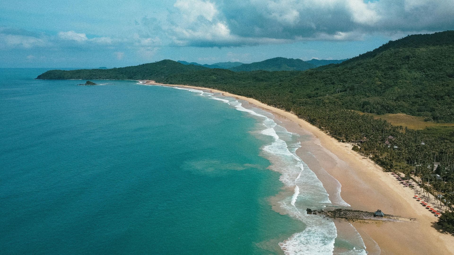 Aerial view of a tropical beach with turquoise water, white sand, and lush green trees. A dark object is on the shore.
