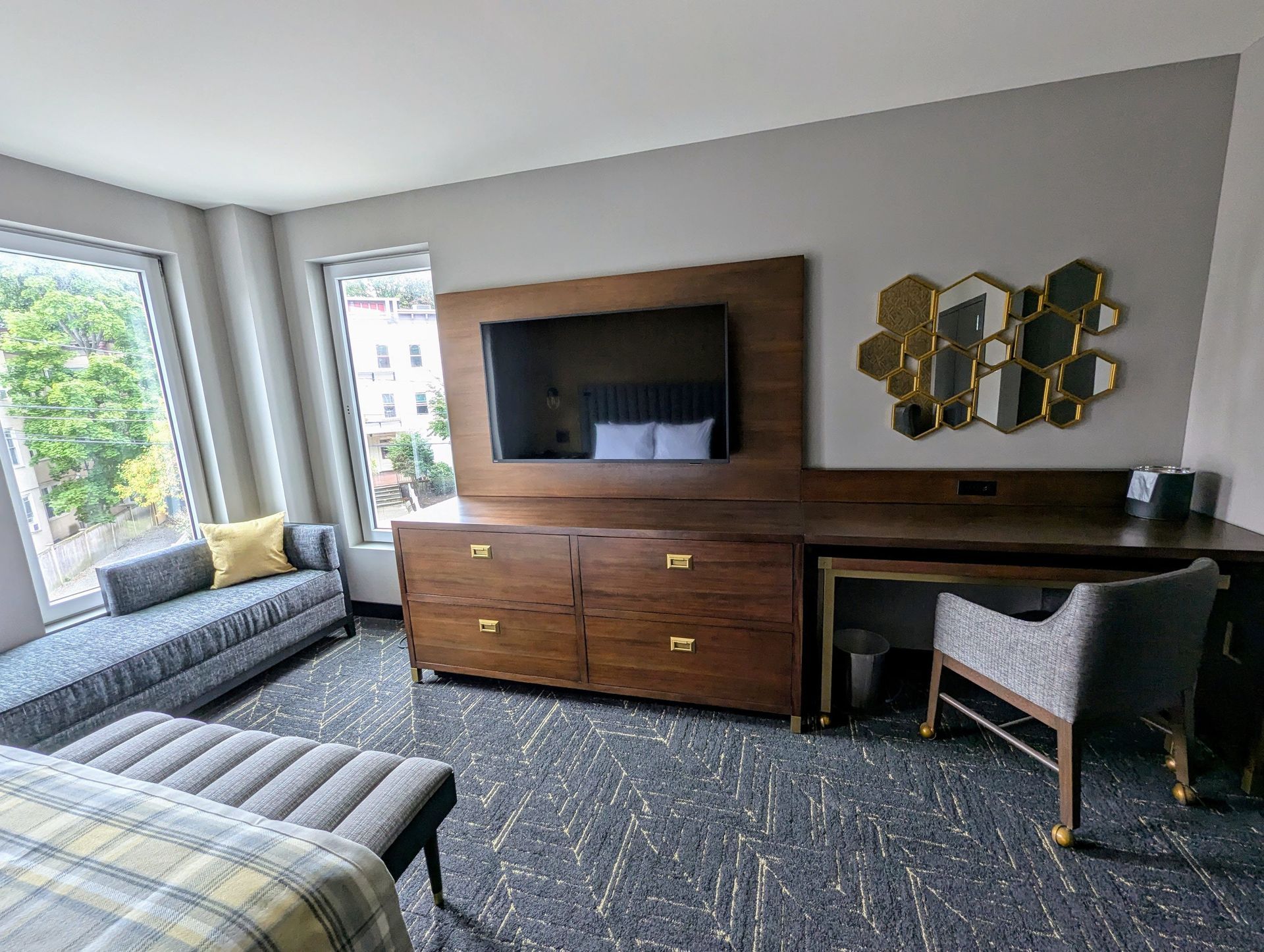 Hotel room with built-in dresser and TV, desk, patterned carpet and bench with a large window.