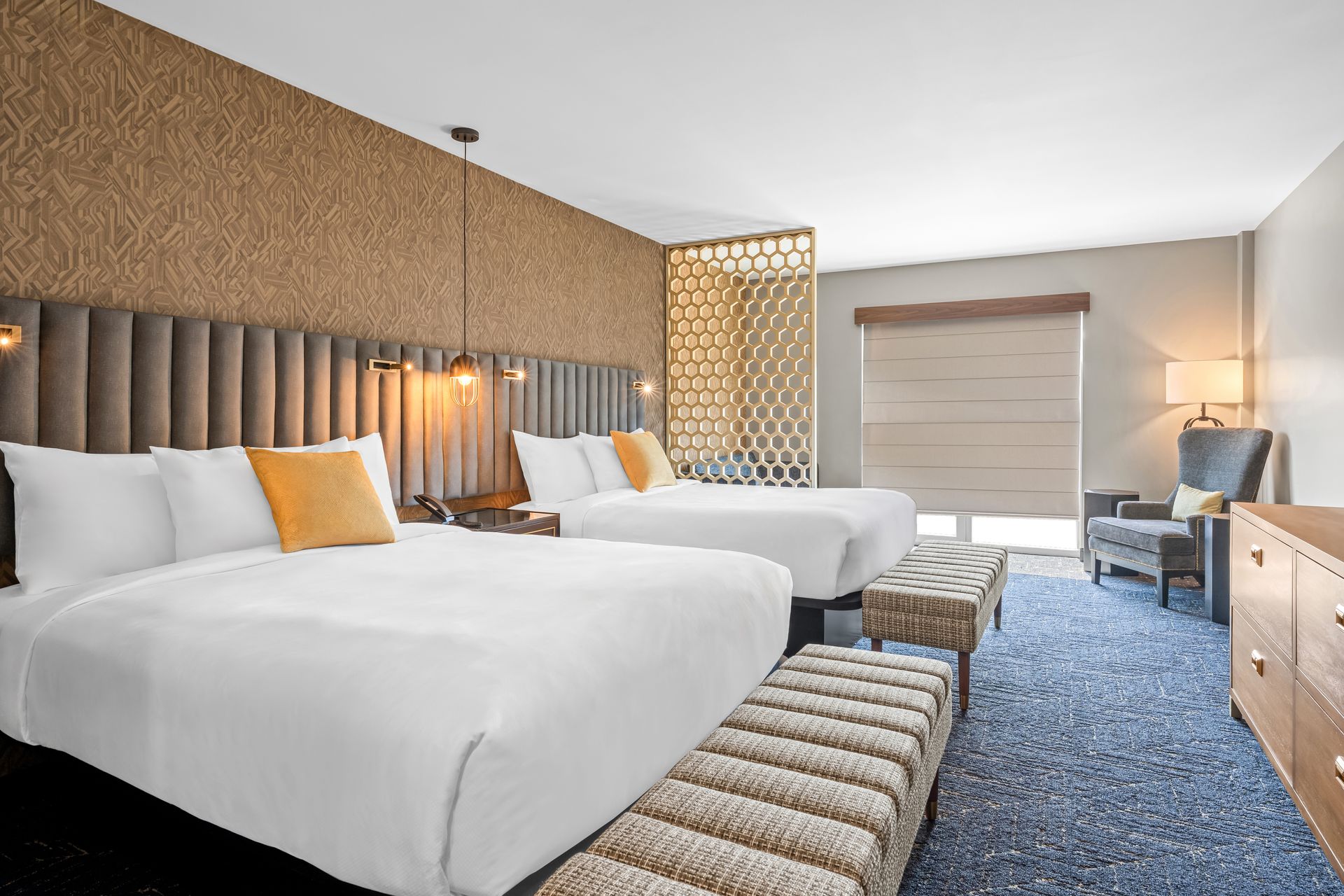 Modern hotel room with two queen beds, blue carpet, wooden furniture, and a window seat