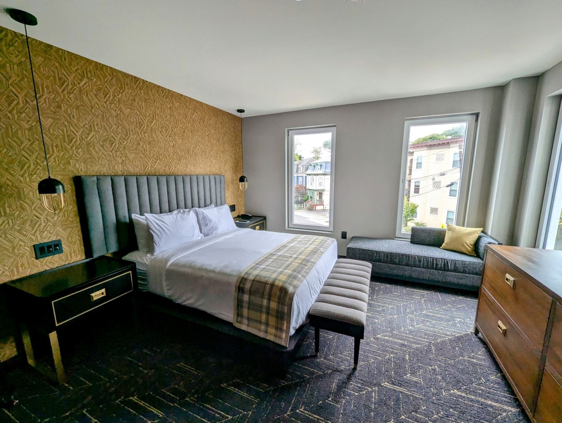 Hotel bedroom with a bed, bench, couch, and dresser. Neutral tones, textured wall.