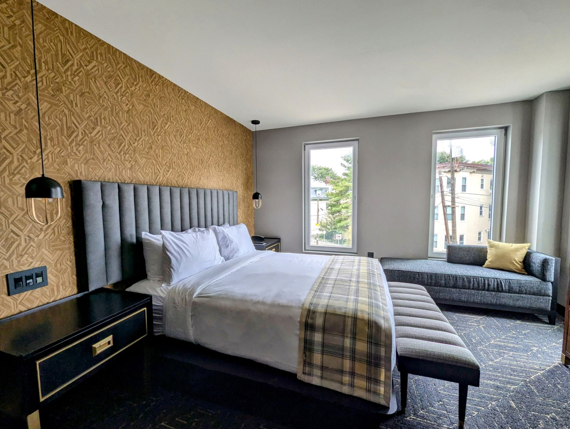 Hotel bedroom with patterned gold wall, large bed, and a grey seating area by windows.
