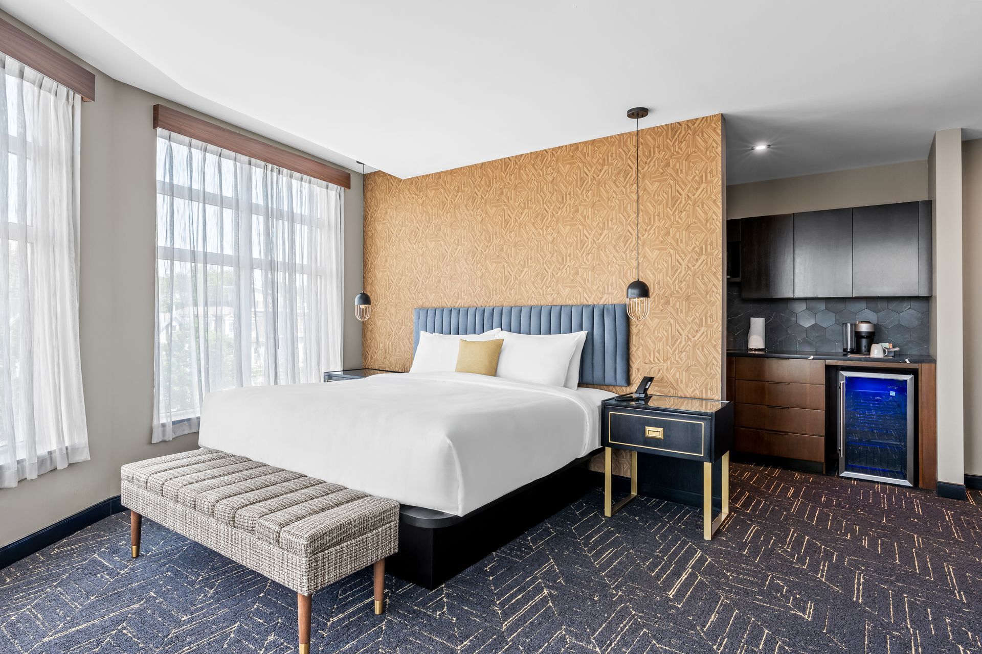 Modern hotel room with king bed, patterned carpet, large windows, desk, and kitchenette with mini fridge