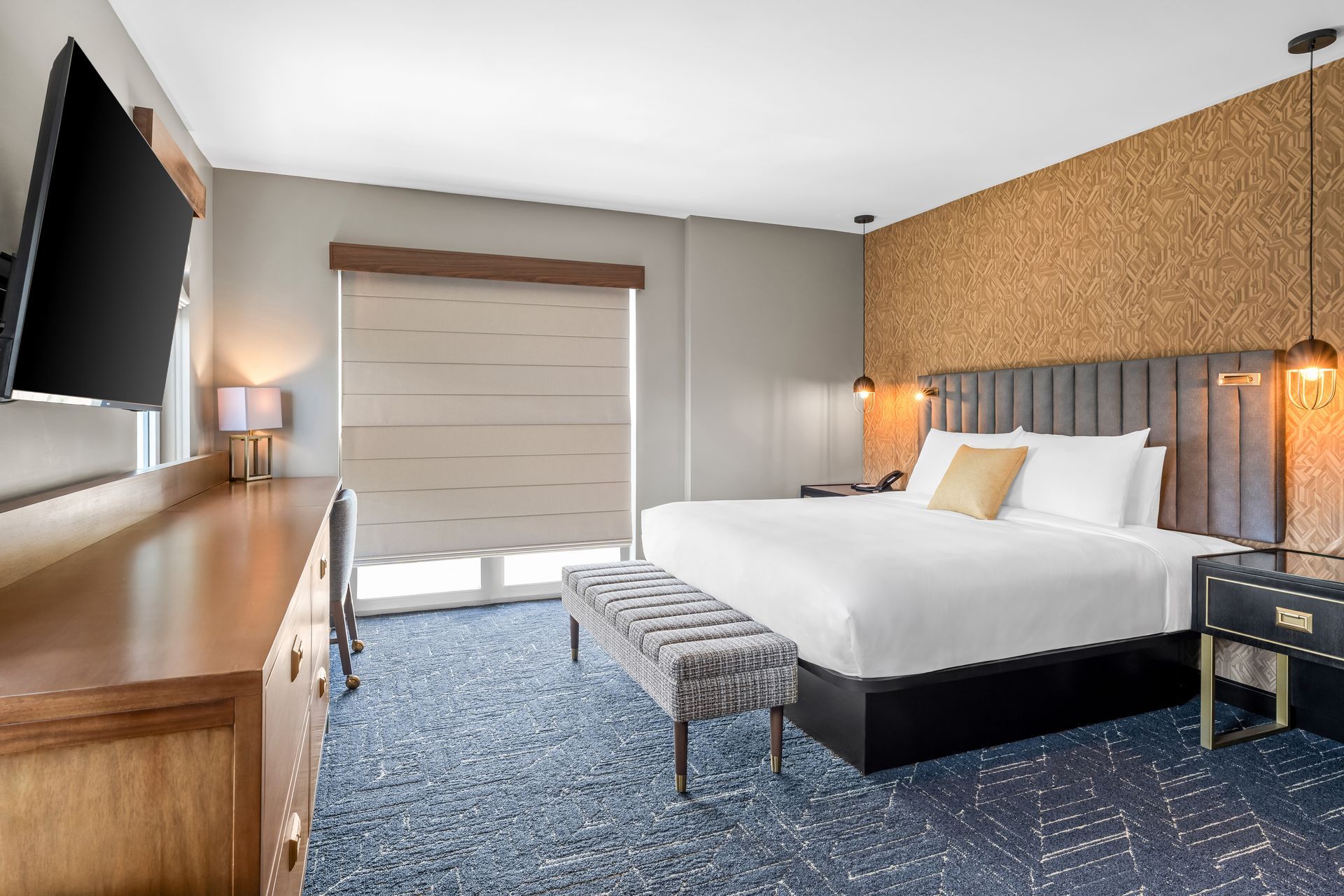 Modern hotel room with a bed, bench, TV, dresser, lamps, and blue carpet.