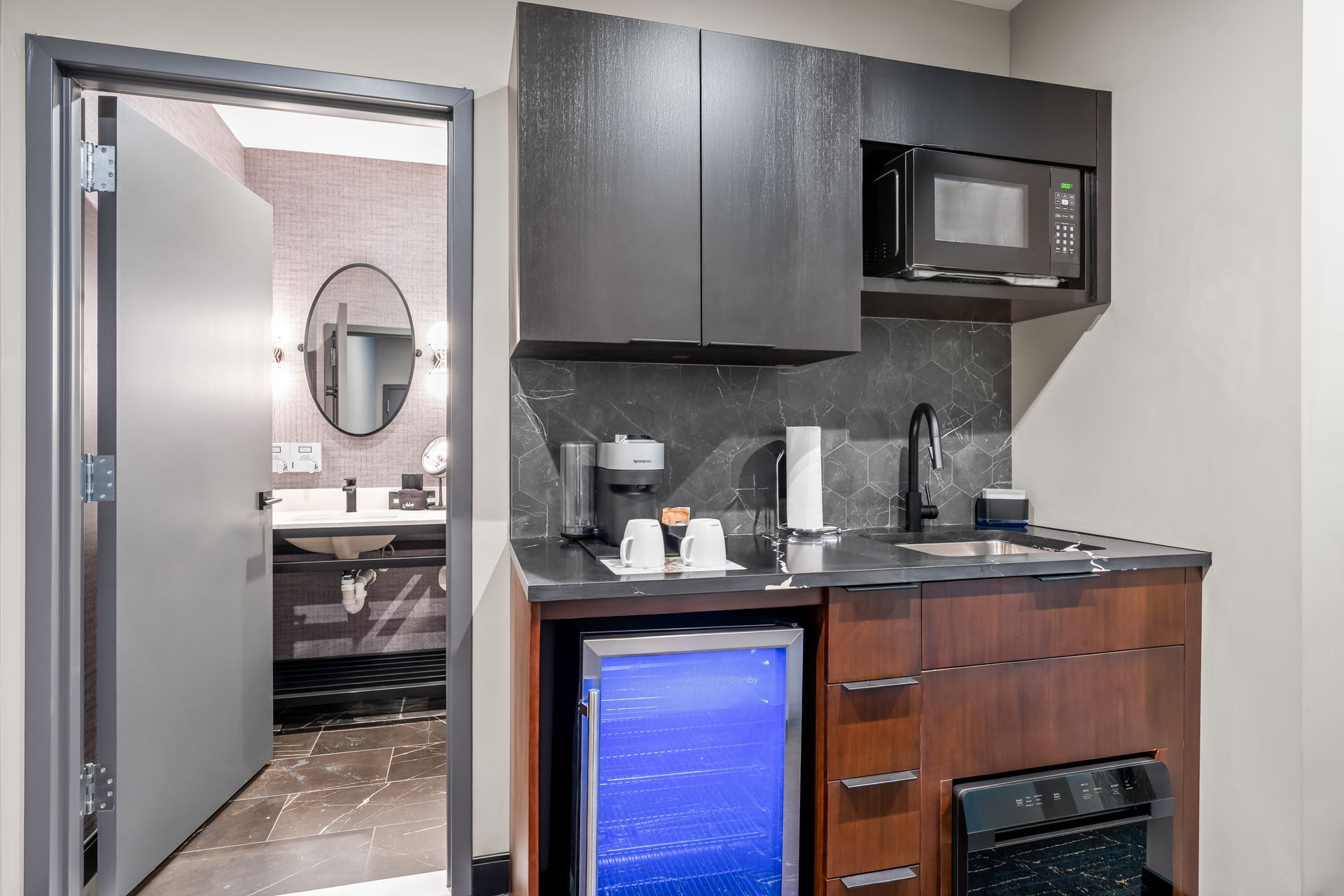 Small kitchenette with dark cabinets and sink beside an open doorway to a bathroom with a round mirror.