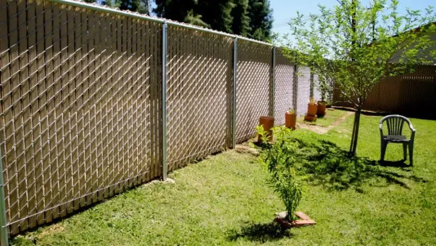 A chain link fence in a backyard with a chair and a tree.