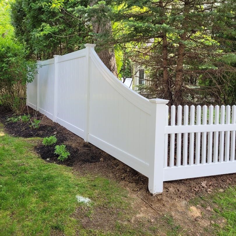 A white picket fence is surrounded by trees in a backyard.