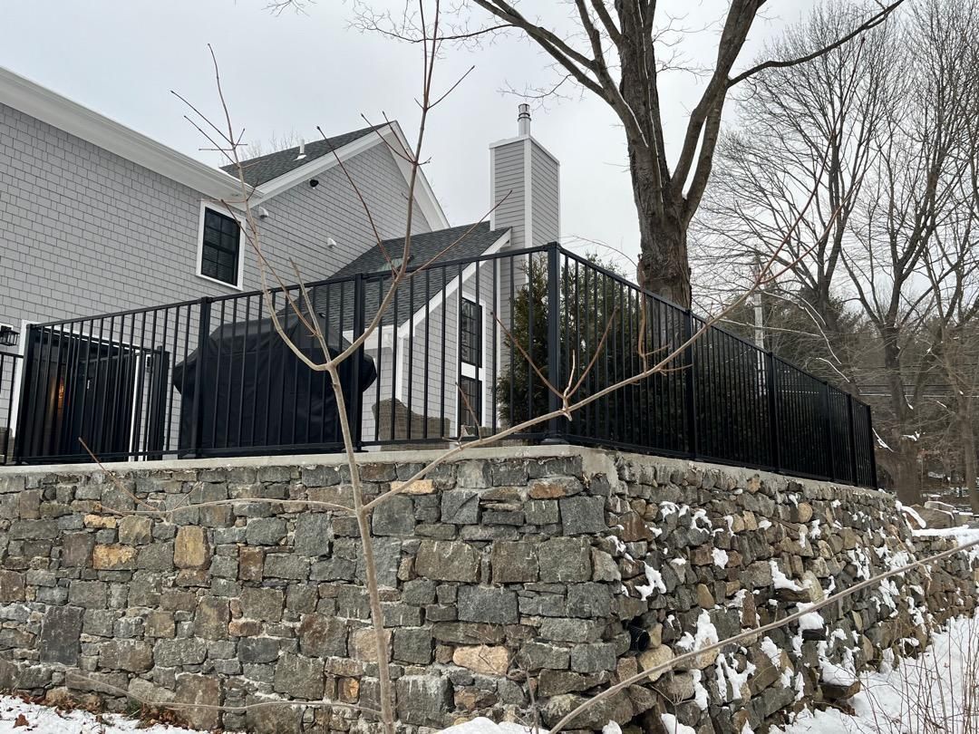 A stone retaining wall topped with a black metal fence in front of a house on a snowy day.