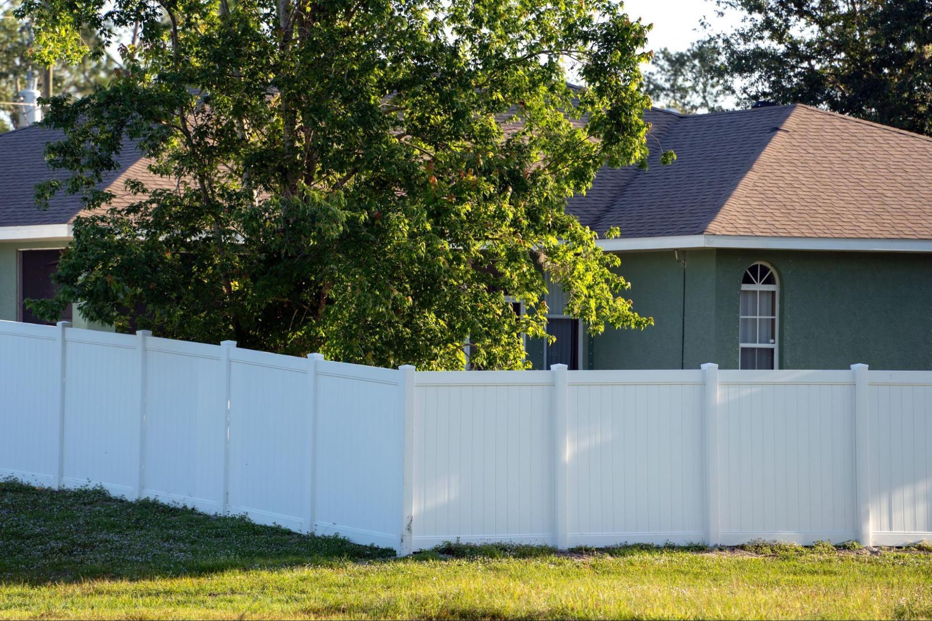 A white vinyl privacy fence sits in front of a green house with a dark roof and a large tree, all under a bright sun.