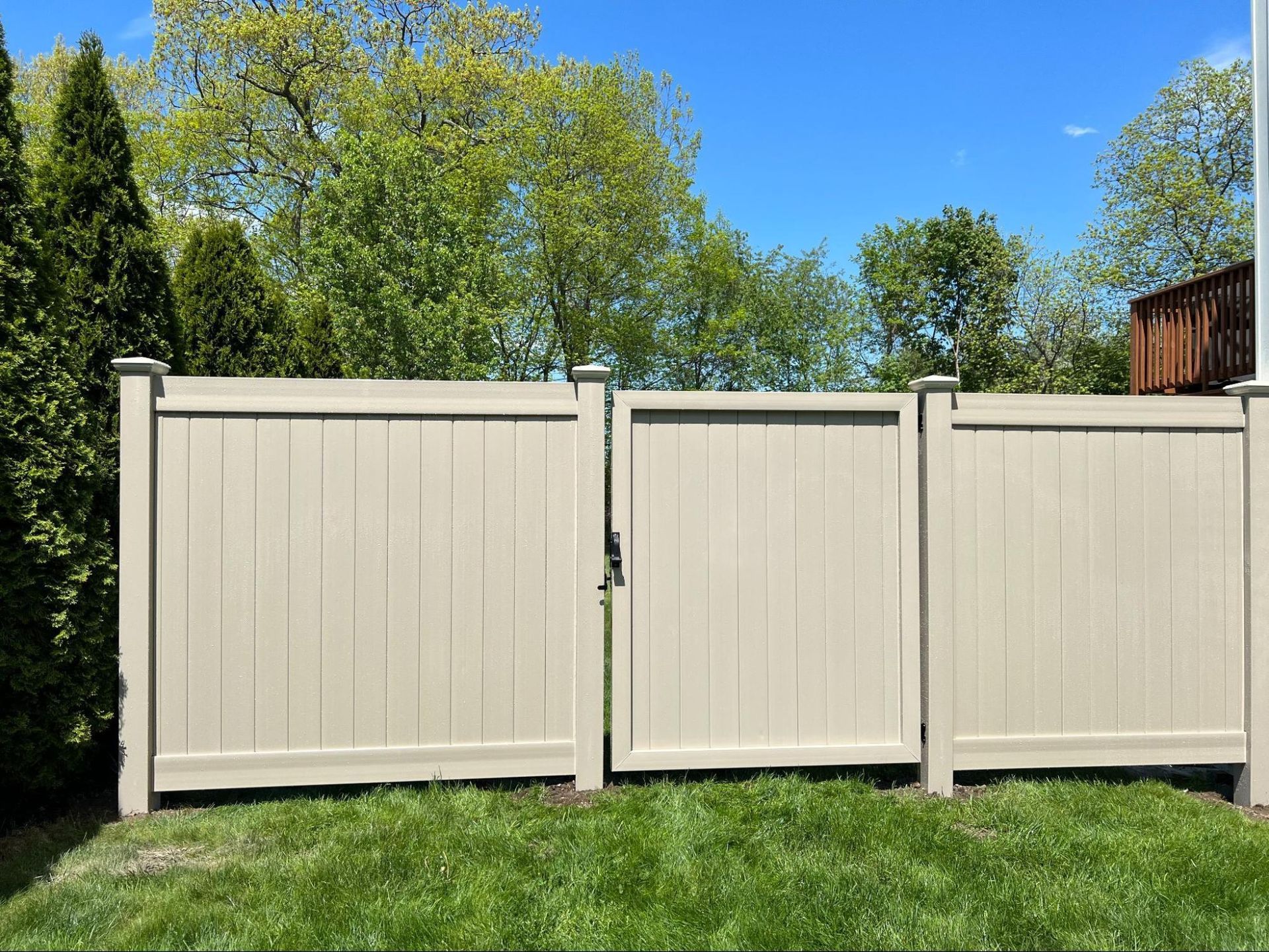 A tan privacy fence with a central gate stands in a grassy backyard, framed by green trees against a clear blue sky.