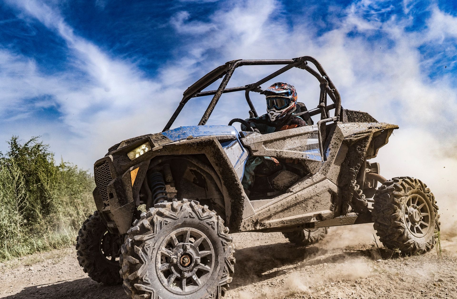 Off-road vehicle speeding on a dirt track, kicking up dust. Driver in helmet, blue and grey vehicle.