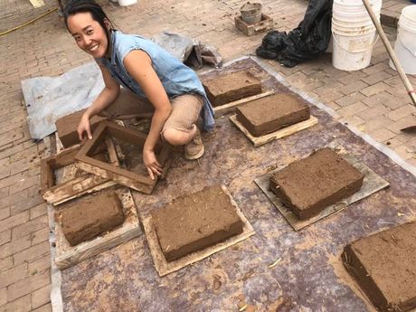 Alice Ko sitting on a brick sidewalk surrounded by adobe bricks she just made, holding an adobe brick form