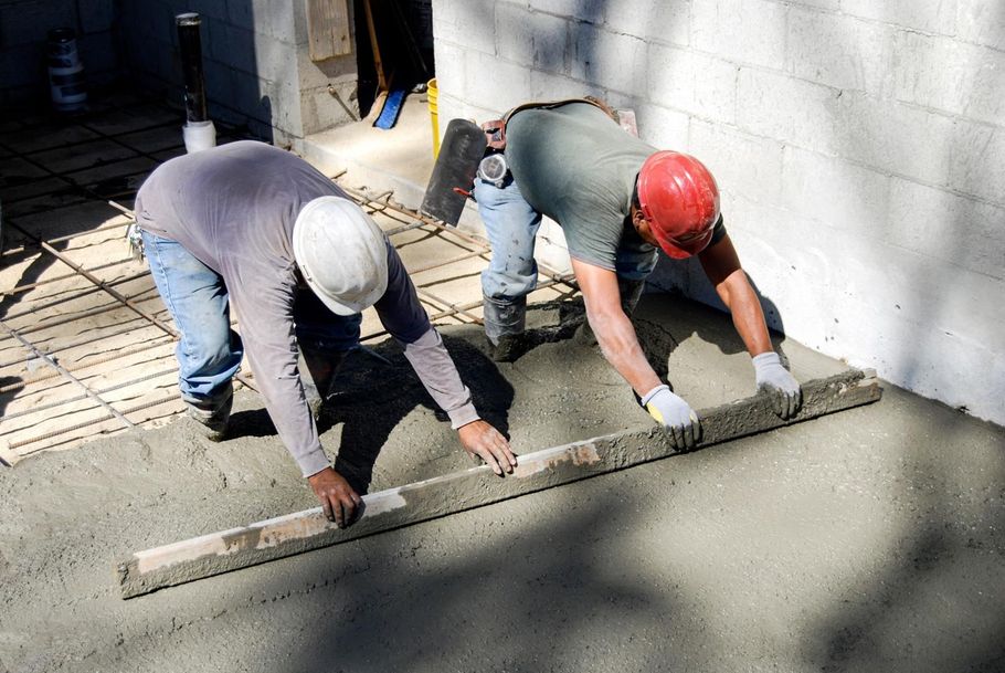 Two workers in hard hats leveling wet concrete beside a cinder block wall.