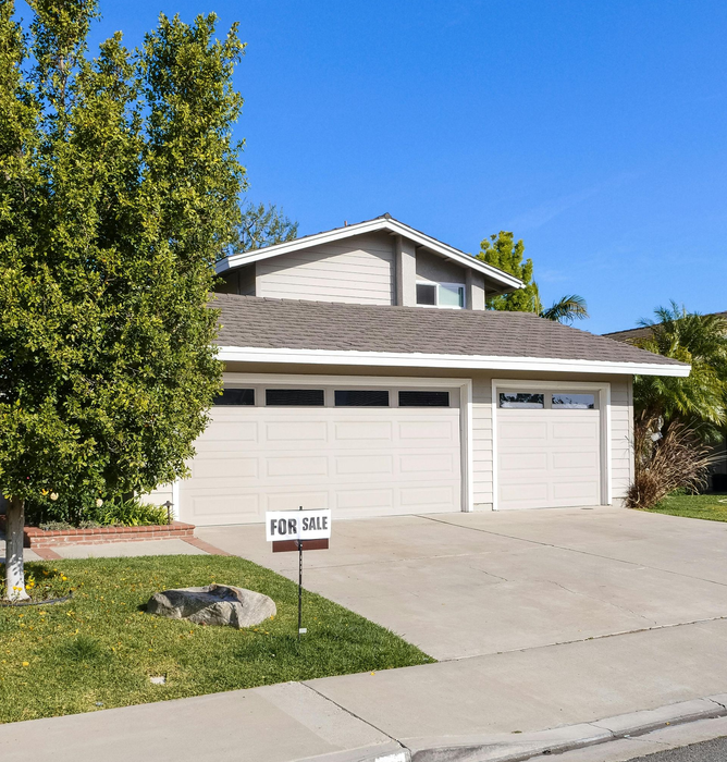 Suburban two-story house with white garage doors, driveway, and trees under a clear blue sky