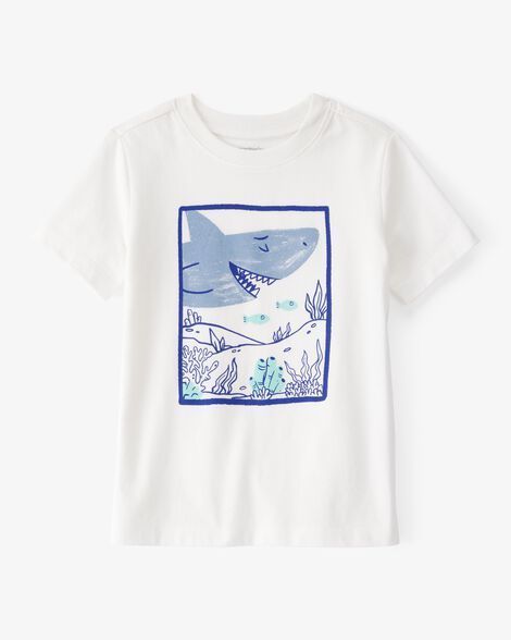 Camiseta blanca con un tiburón azul dentro de un cuadrado azul. El tiburón tiene la boca abierta y está rodeado de plantas submarinas.
