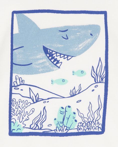 Camiseta blanca con un tiburón azul dentro de un cuadrado azul. El tiburón tiene la boca abierta y está rodeado de plantas submarinas.