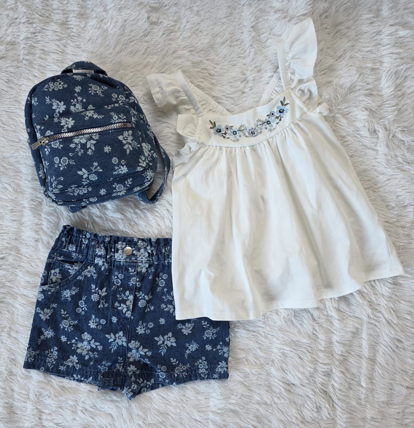 Un conjunto de niño: camiseta blanca con bordado azul, pantalones cortos de mezclilla y una mochila a juego, todo sobre una superficie blanca esponjosa.