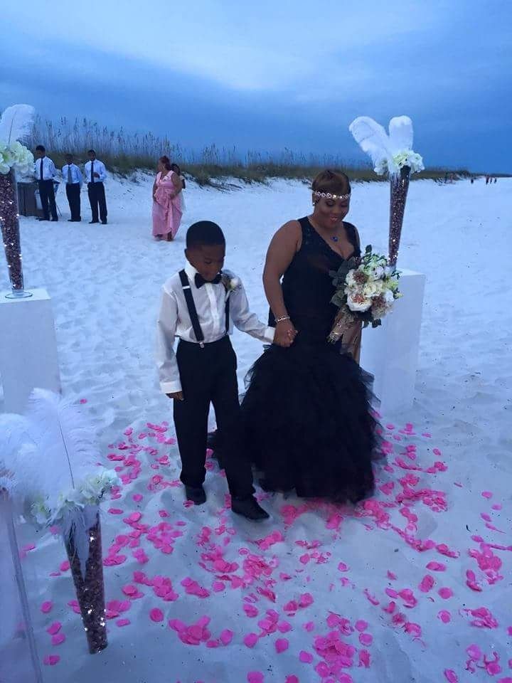 Couple in formal attire posing on a rose-petal-strewn beach at dusk, with guests and white decorations nearby