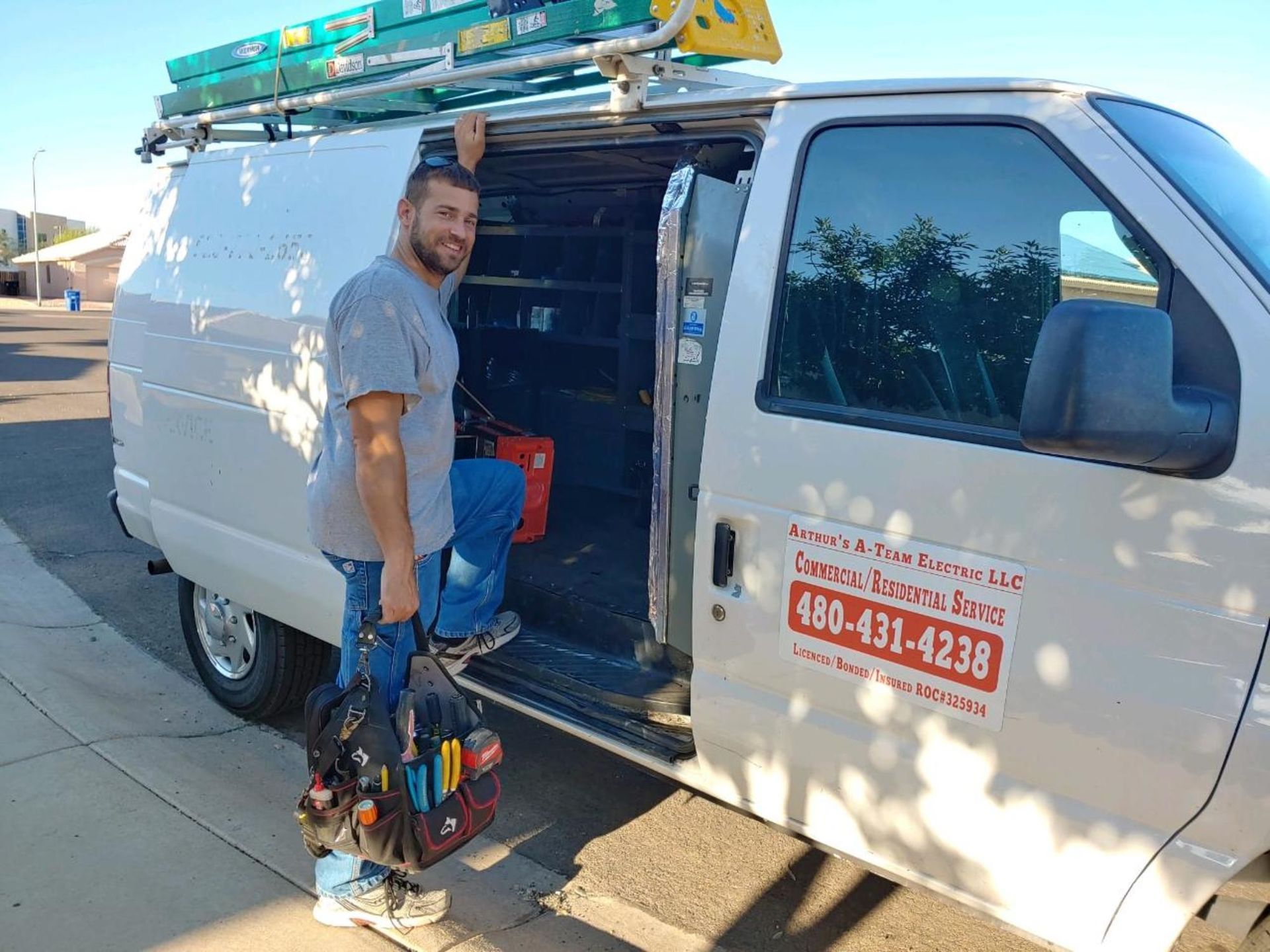 A man is standing next to a white van with a ladder on top of it.