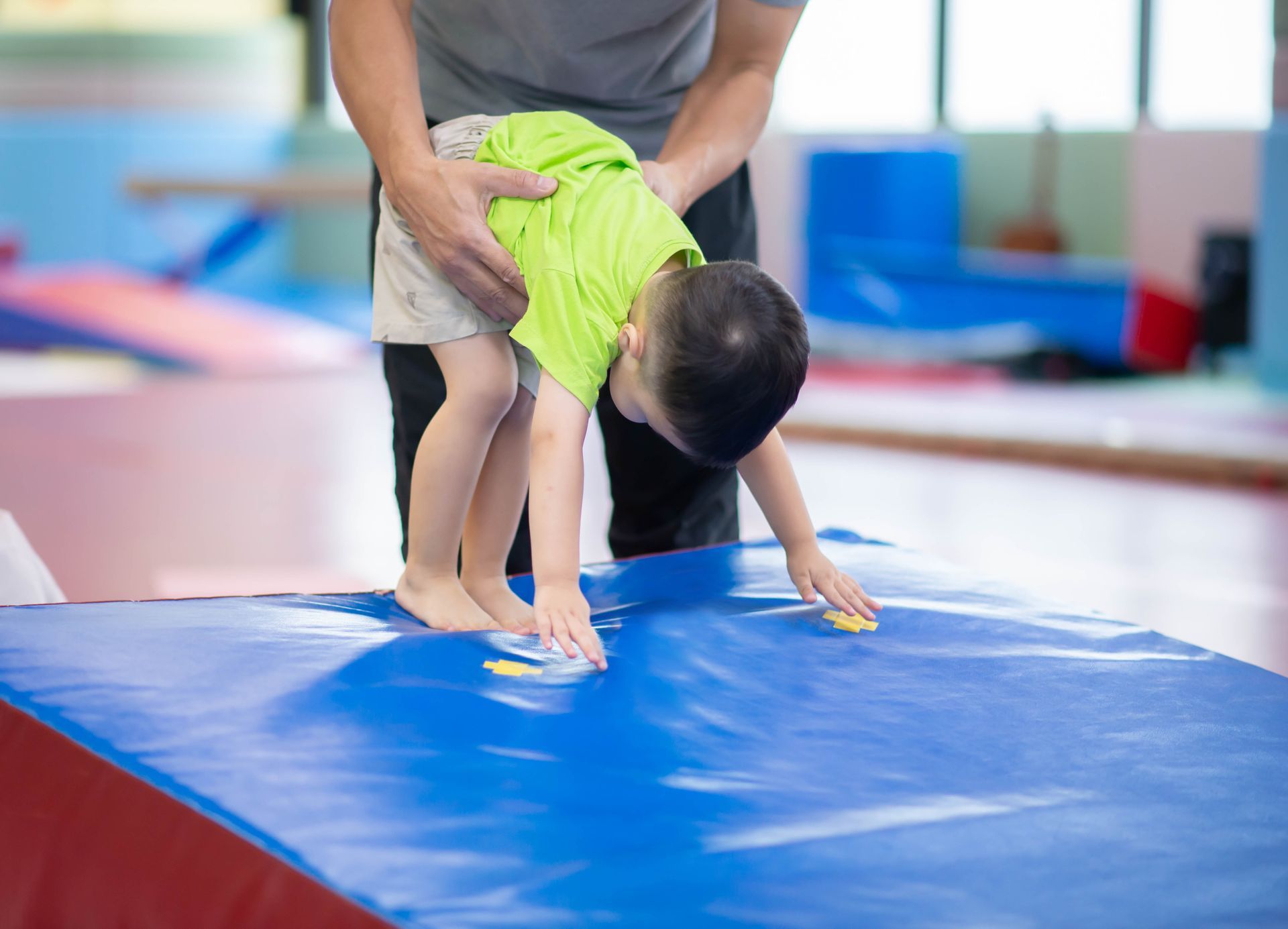 Woman helps a child balance on a wooden beam in a gym; he has arms out, smiling.