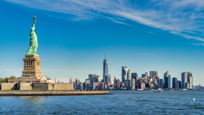 The statue of liberty is surrounded by water and a city skyline in the background.