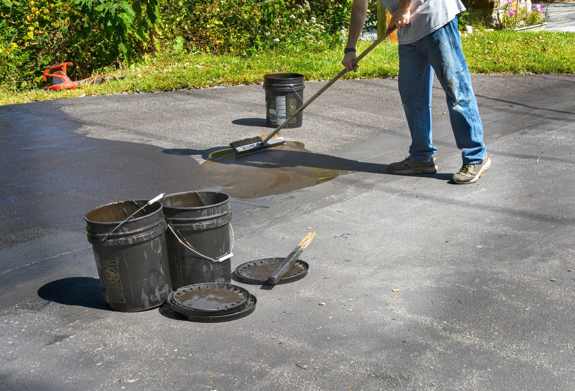 Person applying black coating to a driveway with paint buckets nearby