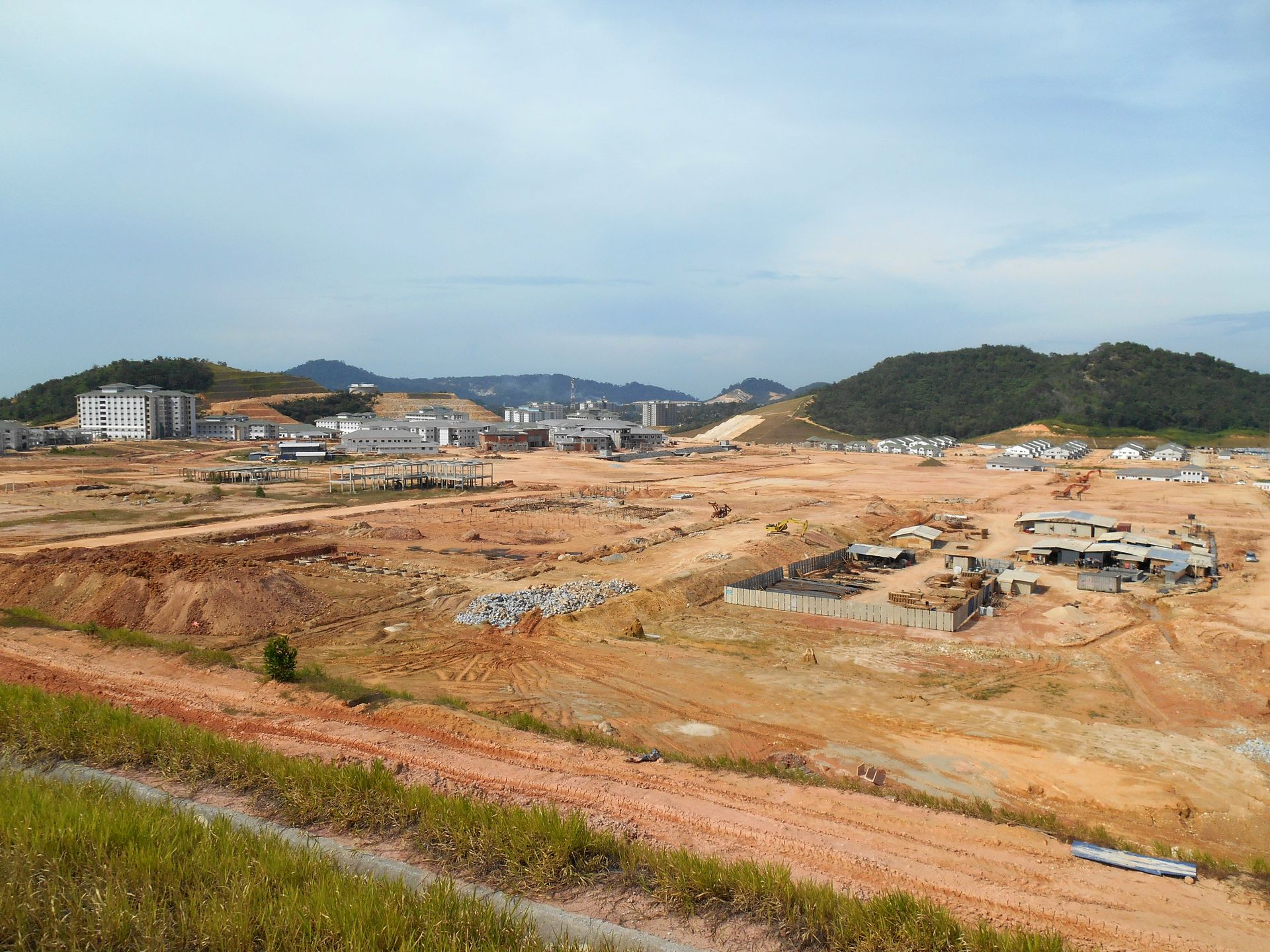 Wide construction site with bare red earth, scattered buildings, and green hills under a cloudy sky