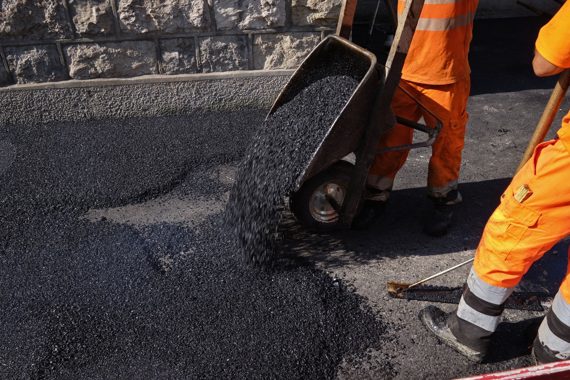 Workers in orange uniforms paving a road with black asphalt near a stone wall
