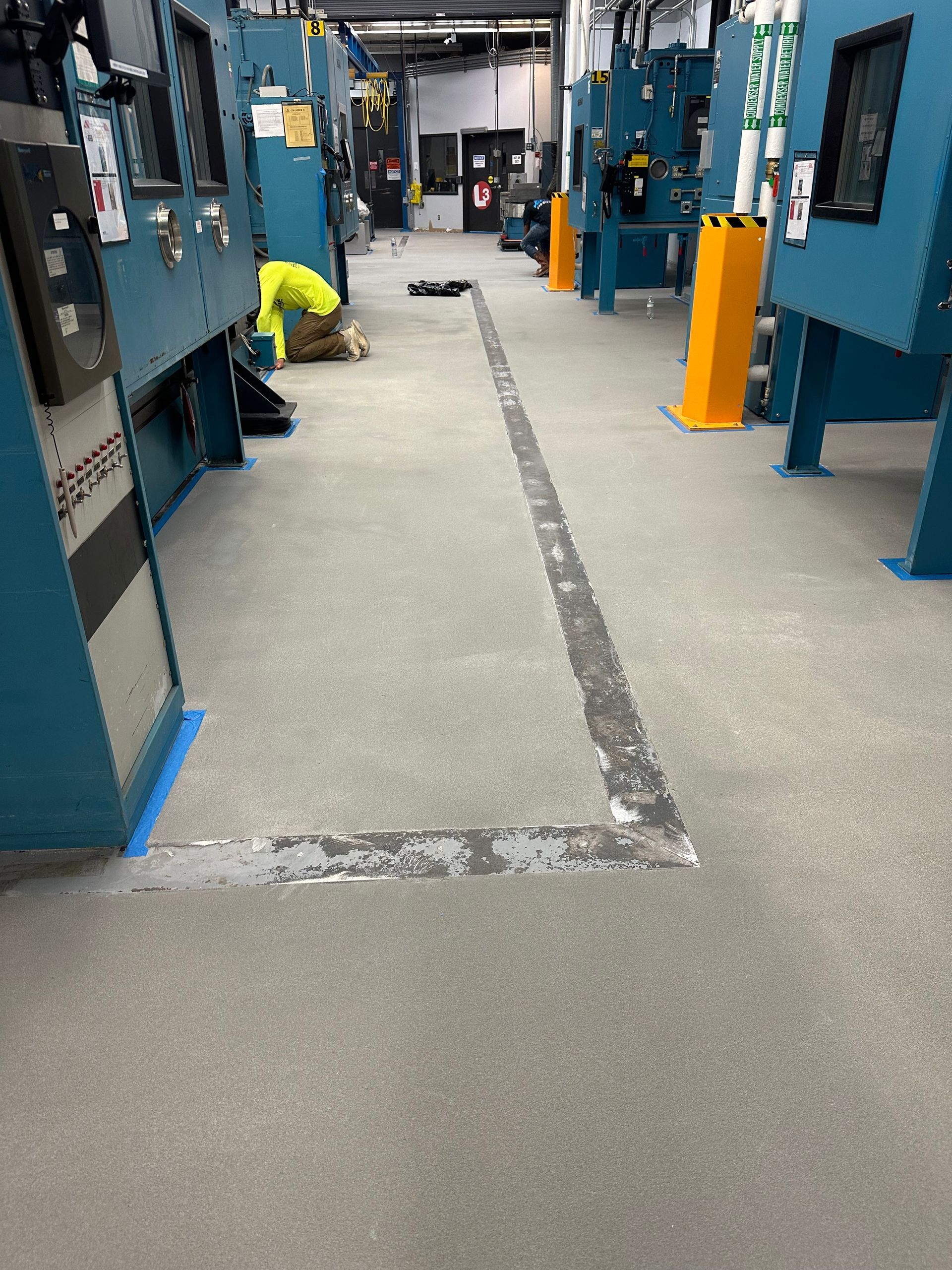 Industrial room with blue machines and a yellow bin along a taped walkway on a gray floor.