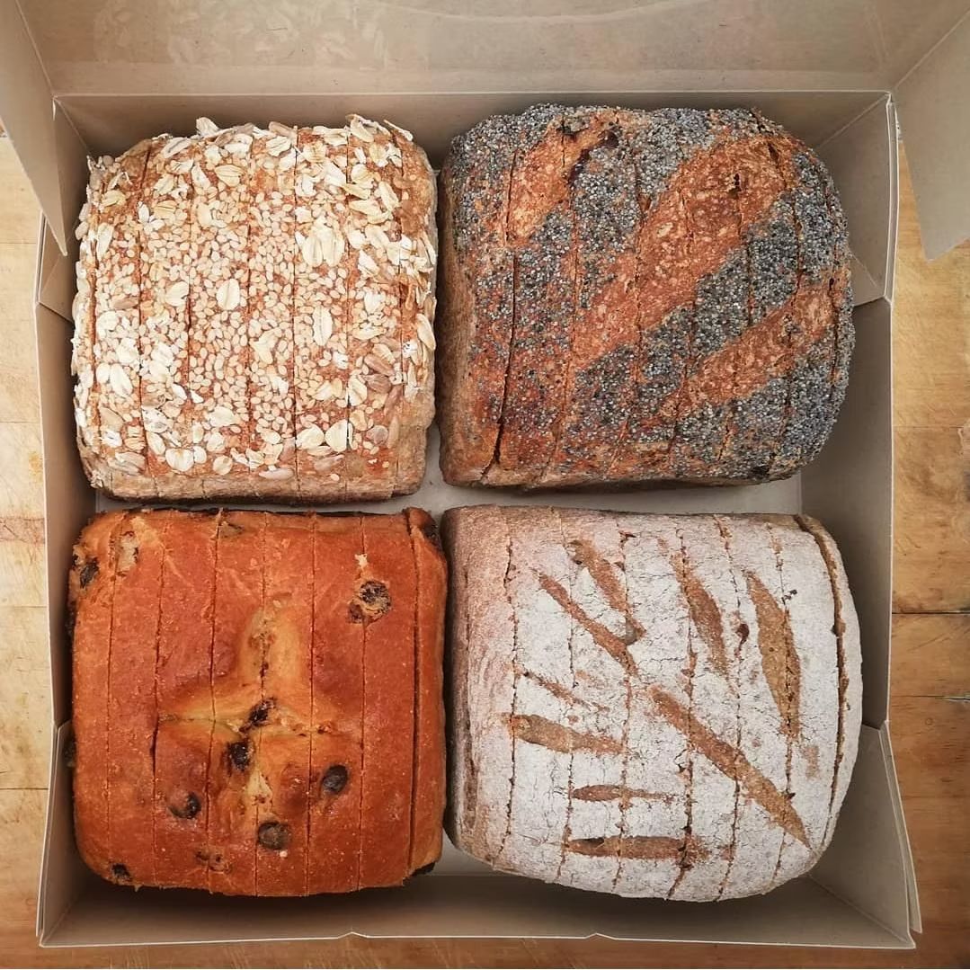 Cuatro panes cuadrados de pan artesanal en una caja; con cobertura de avena, semillas, pasas y diseños rayados.