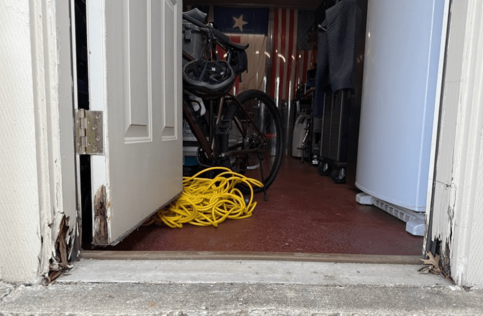 Open doorway to cluttered storage space; yellow cord, bike, and flag are visible.