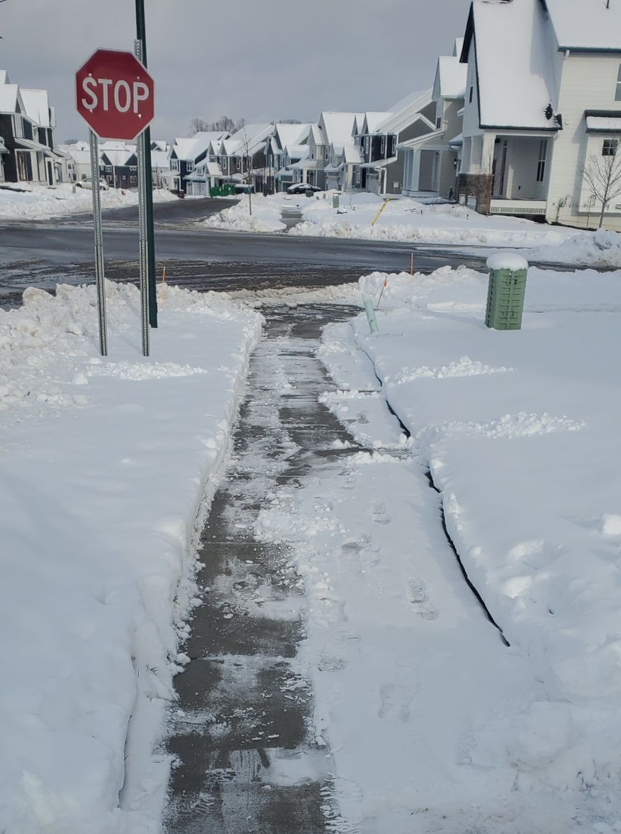 Snow-covered neighborhood street and sidewalk; Stop sign; cleared path through snow; houses in background.