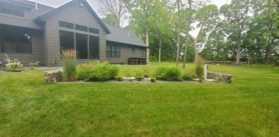 A house with a lush green lawn and garden bed in front of it. Trees are in the background.