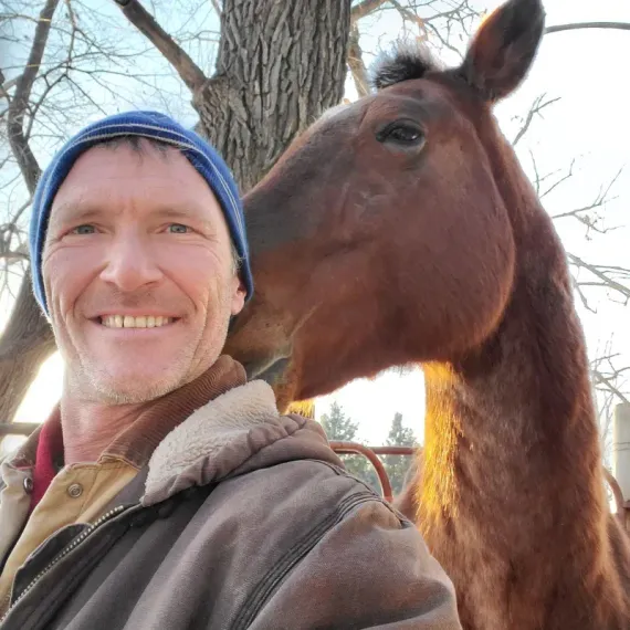 Man smiles as a brown horse nuzzles his face. Outdoors, tree and fence in background.