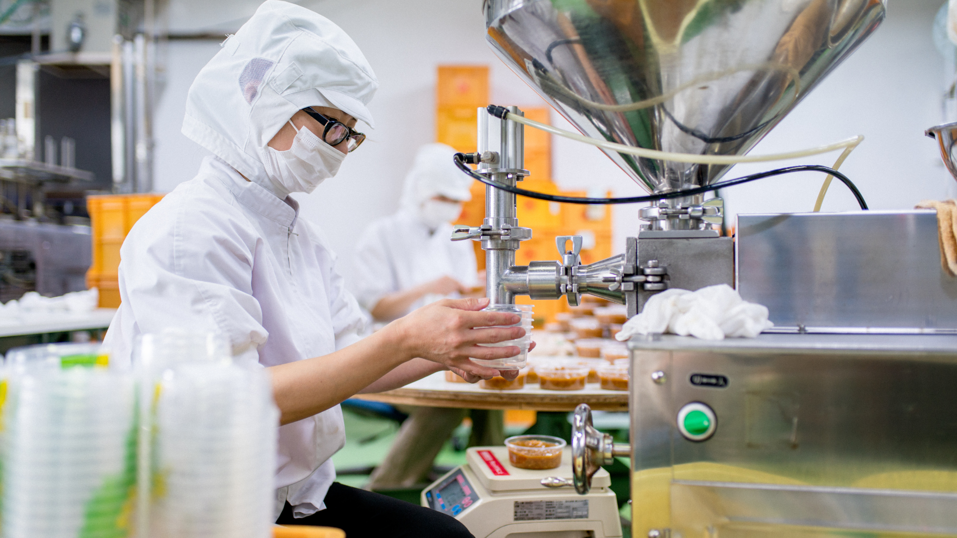 Woman in hair net and mask fills cups at a food processing facility.