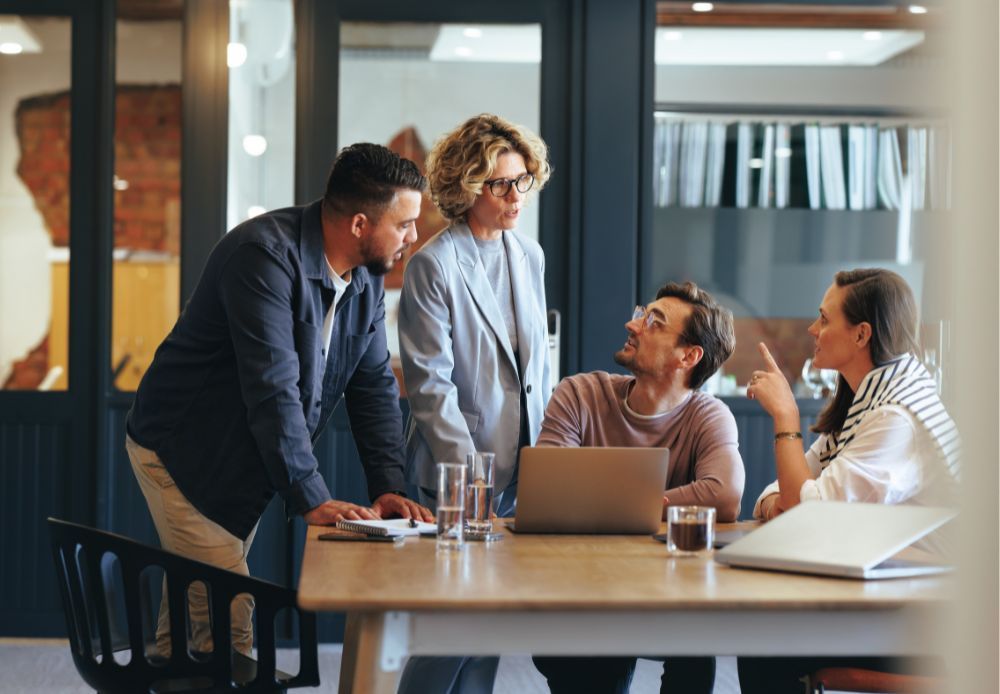 A group of people are standing around a table with a laptop.