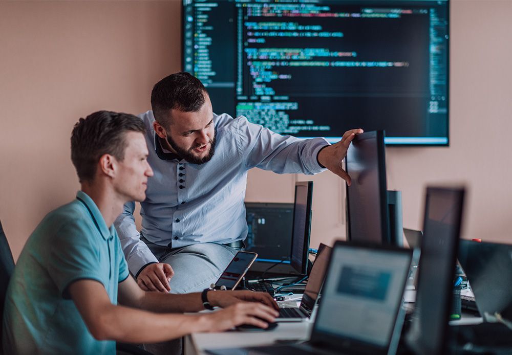 A man is pointing at a computer screen while another man looks on.