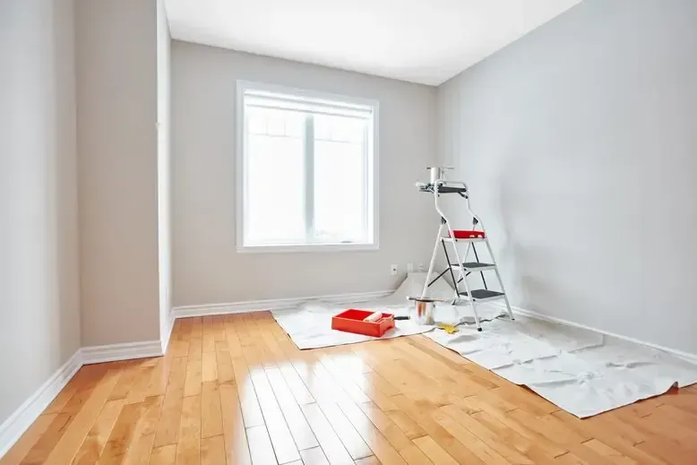 Empty room with hardwood floor, freshly painted gray walls, painting supplies by a window.
