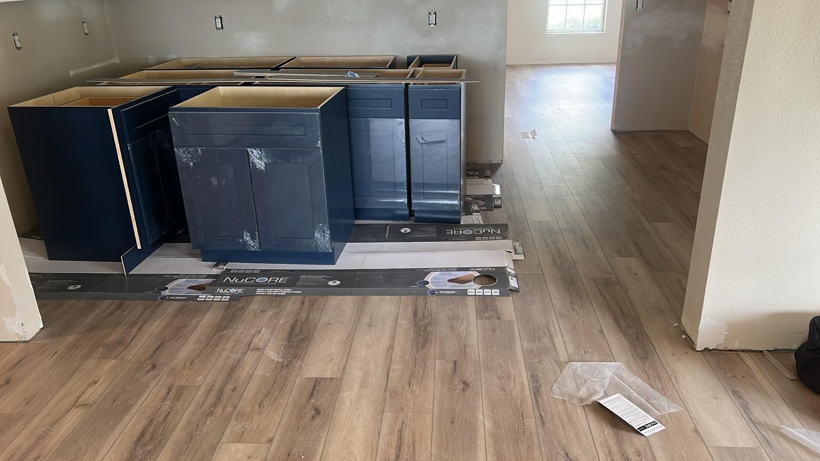 Blue kitchen cabinets on cardboard in a room with wood-look flooring and a hallway.